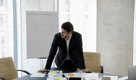 Corporate man in black suit leaning on his desk, in an office with a flip chart in the background.